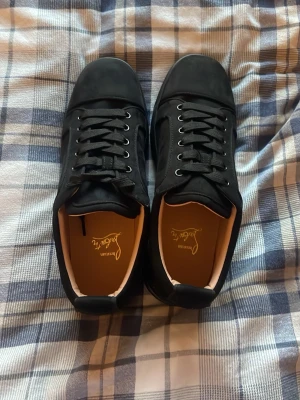 Svarta sneakers från Christian Louboutin - Stilrena svarta sneakers från Christian Louboutin med lågt skaft, svarta skosnören och diskret logga på hälen. Skorna har ovandel i mocka och detaljer i läder, samt klassisk gummisula. Perfekta för dig som gillar exklusiv och clean streetstyle.