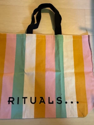 Randig shoppingväska från Rituals - Färgglad shoppingväska från Rituals med breda ränder i pastellfärger som rosa, mintgrön, gul och vit. Väskan har svarta handtag och texten 'SMILE AND THE WORLD SMILES BACK' på ena sidan. Tillverkad i tyg, perfekt för shopping eller stranden.