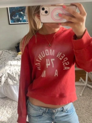 Röd sweatshirt med tryck - Säljer en röd sweatshirt med vitt tryck på bröstet där det står 'ASPEN MOUNTAIN'. Tröjan har en avslappnad passform, rund halsringning och långa ärmar. Perfekt till jeans för en casual look.