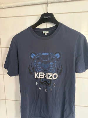 Blå Kenzo t-shirt  - Säljer en blå t-shirt från Kenzo med det ikoniska tigertrycket och texten 'KENZO PARIS' på bröstet. T-shirten har rund hals och korta ärmar. Storlek S, hör av er vid frågor. 
