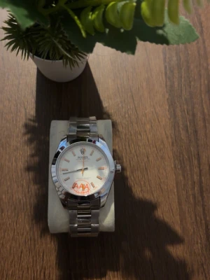 Klocka  - Stilren klocka i silverfärg med metallarmband och vit urtavla. Indexmarkeringar och visare i guldton ger en snygg kontrast. Klassisk rund form och enkel design som passar till många stilar.