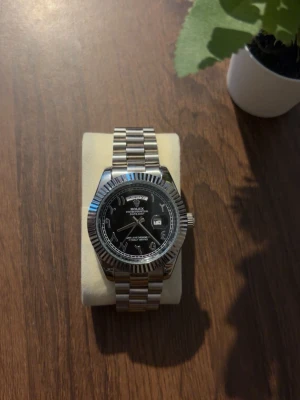 Klocka  - Stilren klocka med silverfärgad länkarmband och svart urtavla. Klockan har datumvisning och tydliga markeringar. Klassisk design med räfflad bezel och robust metallarmband.