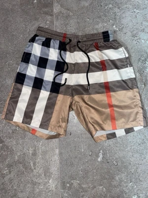 Rutiga badbyxor från Burberry - Säljer ett par snygga badbyxor från Burberry med klassiskt rutmönster i beige, svart, vitt och rött. De har elastisk midja med svart dragsko och meshfoder på insidan. Perfekta för stranden eller poolen!