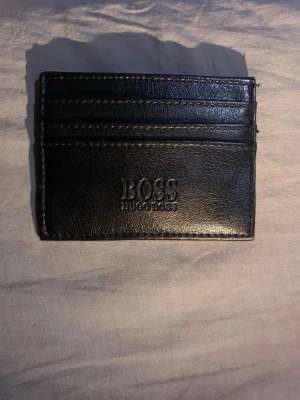 Svart kortfodral från Hugo Boss - Snyggt svart kortfodral från Hugo Boss i äkta läder. Fodralet har flera kortfack på båda sidor och en stilren präglad logga på framsidan. Perfekt för dig som vill ha ett smidigt och elegant sätt att bära dina kort.