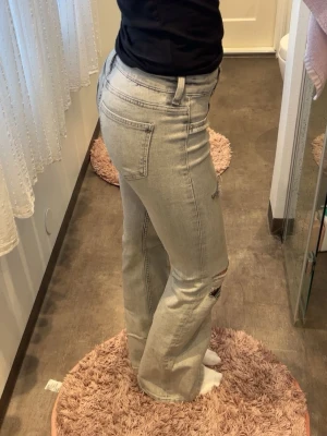Ljusgråa bootcut jeans med slitningar - Säljer ett par ljusgråa jeans med bootcut passform och slitningar vid knäna. Jeansen har klassiska fem fickor och hög midja. Perfekta för dig som gillar en avslappnad men trendig stil.