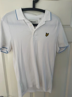 Vit Lyle Scott pikétröja  - Klassisk vit pikétröja från Lyle & Scott med blå ränder på krage och ärmslut. Tröjan har korta ärmar, knappslå framtill och den ikoniska gula loggan broderad på bröstet. Perfekt till sommaren för en clean och sportig look. Kontakta för frågor eller förhandling!