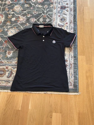 Moncler pike - moncler Klassisk svart pikétröja med korta ärmar och krage. Tröjan har vita och röda detaljer på krage och ärmslut samt knappslå framtill. Liten logotyp på bröstet ger en sportig touch. Perfekt till jeans eller kjol för en avslappnad look.