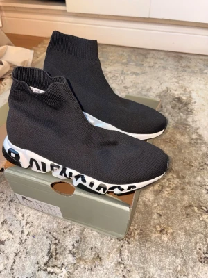 Svarta sock sneakers med grafisk sula - Säljer ett par svarta sock sneakers med stretchig stickad ovandel och vit sula med svart grafiskt mönster. Skorna har en strumpliknande passform och ribbad kant upptill. Perfekta för dig som gillar en modern och clean stil.