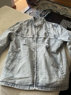 Ljusgrå distressed jeansjacka - Säljer en ljusgrå jeansjacka med dragkedja och ståkrage. Jackan har slitna detaljer på bröstet och magen samt två snedställda fickor framtill. Perfekt för dig som gillar en streetstyle-look med lite edge.