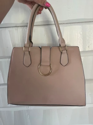 Beige handväska med gulddetalj - Stilren beige handväska i slätt konstläder med två rejäla handtag och en snygg guldfärgad ringdetalj framtill. Väskan har en klassisk fyrkantig form och svarta kanter som kontrast. Perfekt för dig som gillar enkel men trendig design.