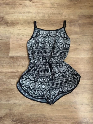 Svartvit mönstrad jumpsuit med smala axelband - Snygg svartvit jumpsuit med grafiskt mönster och smala axelband. Modellen har resår i midjan och knytband för extra passform. Lätt och luftig, perfekt för varma dagar. Mjuk och bekväm känsla med korta ben.