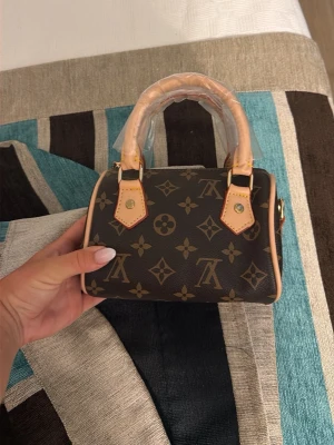 Brun monogram handväska - Snygg liten handväska i brun färg med klassiskt monogrammönster och beige detaljer. Väskan har två korta handtag och guldfärgade metalldetaljer. Perfekt för dig som gillar trendiga accessoarer med lyxig känsla. Lite färg som har kommit på några delar men detta kan man ta bort enkelt själv 