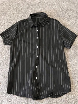 Svart kortärmad randig skjorta - Snygg svart skjorta med vita vertikala ränder och korta ärmar. Skjortan har klassisk krage och vita knappar framtill. Perfekt för en stilren och avslappnad look.