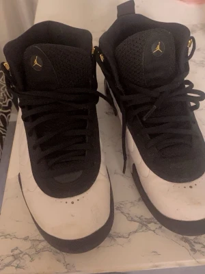 Svarta och vita Air Jordan 12 sneakers - Snygga Air Jordan 12 sneakers i svart och vitt med gulddetaljer. Skorna har högt skaft, klassisk Jumpman-logga på sidan och tungan samt vågigt mönster på sidorna. Ovandelen är i mocka och läder, med svarta skosnören och robust sula. Stl 42.5