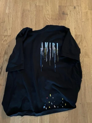 Svart AMIRI t-shirt med färgstänk - Snygg svart t-shirt från AMIRI med färgstänk och droppande logga på bröstet. T-shirten har korta ärmar och en klassisk passform. Perfekt för dig som gillar streetwear och vill sticka ut med coola detaljer. Pris kan diskuteras vid snabb affär! Bli inte lurrade det står xl men den är L 