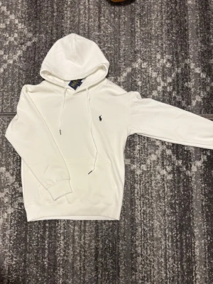 Vit hoodie från Polo Ralph Lauren - Snygg vit hoodie från Polo Ralph Lauren i 100% bomull. Klassisk design med huva och dragsnören, samt den ikoniska lilla loggan broderad på bröstet. Perfekt för en clean och stilren look.