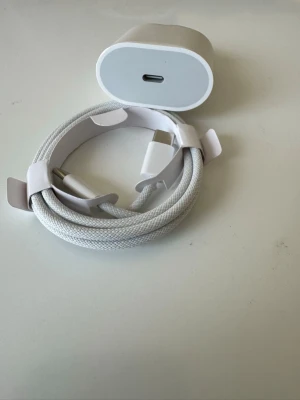 IPhone 17 Vit USB-C laddare och kabel - 20w eladapter  vit USB-C laddare med tillhörande flätad laddningskabel. Kabeln har USB-C kontakter i båda ändar och är tillverkad i slitstarkt textilmaterial. Perfekt för snabbladdning av mobiltelefoner och andra enheter. Enkel och stilren design.