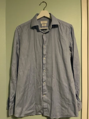 Ljusblå skjorta från Massimo Dutti - Snygg ljusblå skjorta från Massimo Dutti i bomull med diskreta ränder. Klassisk krage och knäppning framtill, perfekt för en clean och stilren look. Långärmad modell med en lätt och bekväm känsla. Strlk 39