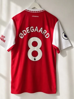 Arsenal 2022/23 Hemmatröja #8 Martin Ødegaard – Storlek L - Arsenal hemmatröja från säsongen 2022/23 med Martin Ødegaard #8 på ryggen. En av de mest uppskattade Arsenal-tröjorna de senaste åren – den klassiska rödvita designen med den ikoniska kragen.  Spelare: Martin Ødegaard Nummer: 8 Klubb: Arsenal FC Säsong: 2022/23 Premier League-märke på ena ärmen Visit Rwanda-sponsor på andra ärmen  Storlek: L  Mått:  Armhåla till armhåla: 55 cm  Längd: 74 cm  