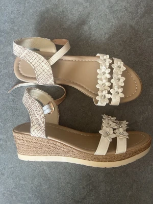 Beige sandal - Säljer ett par beiga sandaler med kilklack. Jättefina vita blommor har den och är justerbart med spänne runt ankeln och mjuk innersula för extra komfort. Perfekta för sommaren och ger en söt touch till din outfit. Helt nya är de, de är aldrig använda men blivit öppna från förpackningen + saknar prislapp