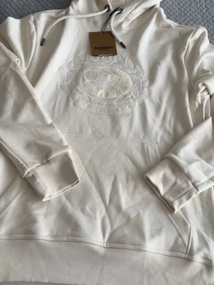 Vit hoodie med broderad Burberry-logga - Säljer en stilren vit hoodie från Burberry med broderad logga och riddarmotiv på bröstet. Hoodien har huva med dragsko och en stor magficka. Tillverkad i mjukt material som känns lyxigt och bekvämt. Perfekt för dig som gillar exklusiva streetwear-plagg.