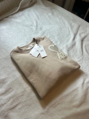 Beige sweatshirt från Ami Paris - Snygg beige sweatshirt från Ami Paris med det ikoniska hjärta-A-märket broderat på bröstet. Tröjan har rund halsringning och ribbade muddar. Perfekt för en clean och stilren look.