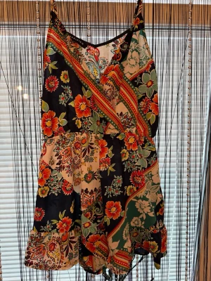 Färgglad blommig jumpsuit med smala axelband - Supersnygg jumpsuit med smala axelband och färgstarkt blommönster i rött, grönt, orange och blått. Lätt och luftig modell med omlott upptill och volangkant nertill. Perfekt för sommardagar