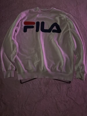 Vit sweatshirt från FILA - Säljer en vit sweatshirt från FILA med stort logotryck i rött och marinblått på bröstet. Tröjan har rund halsringning, långa ärmar och ribbade muddar vid ärmslut och nederkant. Perfekt för en sportig och avslappnad stil.