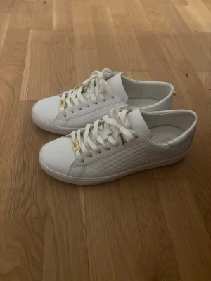 Vita Michael Kors sneakers med gulddetaljer - Snygga vita sneakers från Michael Kors med guldfärgade detaljer på snörningen och diskret MK-logga. Skorna har lågt skaft och ovandel i läder med mönstrad sida. Perfekta för en clean och stilren look. Storlek 40