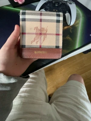 Rutig plånbok från Burberry - Snygg plånbok från Burberry med klassiskt rutigt mönster i beige, svart och lite brunt plånboken  har bruna detaljer och guldfärgad logga. Insidan är mörkbrun med flera kortfack och Burberrys logotyp i guld. Perfekt för dig som gillar stilrena accessoarer.