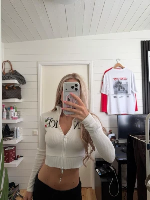 Vit croppad ribbad zip-top - Snygg vit croppad topp med ribbad struktur och dragkedja framtill. Långärmad modell med hög krage och tryckdetaljer på bröstet. Perfekt för en trendig och avslappnad look.