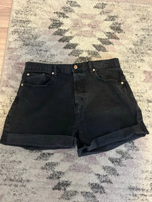 Svarta jeansshorts med hög midja - Svarta jeansshorts med uppvikta benslut. Klassisk femficksmodell med knappgylf och bälteshällor. Snygg och tidlös design! Shortsen är svarta, i storlek XL från Lager 157, ingen prislapp kvar men aldrig använda. Pris kan diskuteras