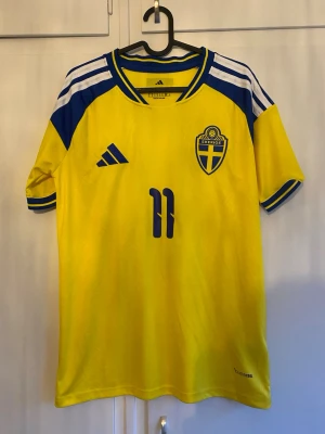 Svensk landslagströja Elanga #11 - Gul och blå Sverige fotbollströja från Adidas med Elanga och nummer 11 på ryggen. Tröjan har korta ärmar, blå detaljer på axlar och ärmslut samt broderad landslagssköld på bröstet. Perfekt för match eller supporterhäng.