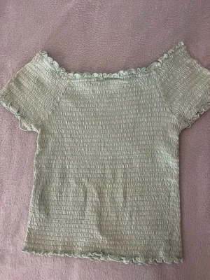 Mintgrön offshoulder topp KappAhl - Superfin mintgrön offshoulder topp från KappAhl med smockad struktur och volangkant vid hals, ärmar och nederkant. Toppens material känns mjukt och stretchigt.