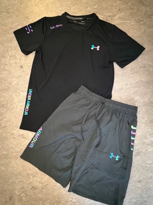 Svart träningsset från Under Armour - Snyggt svart träningsset från Under Armour med t-shirt och shorts. Båda plaggen har färgglada loggor och textdetaljer i regnbågsfärger. Tillverkade i ett lätt och snabbtorkande material som passar perfekt till träning och sportaktiviteter.