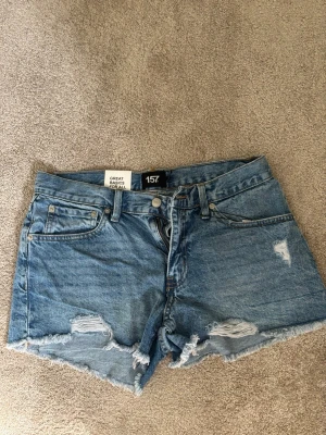 Blå denimshorts med slitningar - Säljer ett par klassiska blå denimshorts från 157 med råa kanter och slitna detaljer framtill. Shortsen har fem fickor, bälteshällor och knappgylf. Perfekta för varma dagar och en avslappnad stil. Shortsen är oanvända med prislappen kvar.