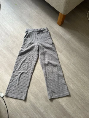 Grå ribbade mjukisbyxor - Säljer ett par grå mjukisbyxor med ribbad struktur och raka ben. Byxorna har resår i midjan och praktiska fickor på sidorna. Perfekta för chill dagar hemma eller när du vill vara bekväm men ändå se snygg ut.