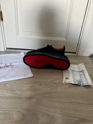 Marinblåa Louboutin sneakers med röd sula - Snygga marinblåa sneakers från Christian Louboutin med den ikoniska röda sulan och diskret logga undertill. Skorna har en klassisk låg siluett och är tillverkade i slätt läder med svarta skosnören. Perfekta för dig som vill sticka ut med stil.