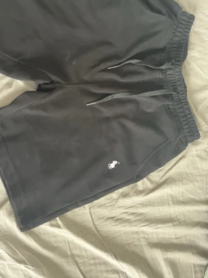 Svarta shorts från Polo Ralph Lauren - Svarta shorts från Polo Ralph Lauren med elastisk midja och dragsko. Klassisk logga broderad i vitt på ena benet. Sköna och stilrena, perfekta för en avslappnad look. Tillverkade i mjukt material för extra komfort.