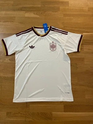 Spanien vm tröja borta - Säljer en vit fotbollströja från Adidas med Spaniens landslagsemblem på bröstet. Tröjan har vinröda detaljer, tre ränder på axlarna och guldkantad krage. Tillverkad i ett glansigt funktionsmaterial, perfekt för träning eller match.   Fraktar samma eler dagen efter du köpt!!! Endast en kvar i lager!!!