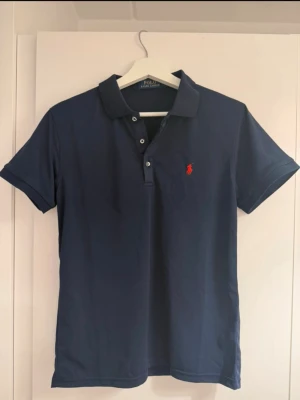 Marinblå pikétröja från Polo Ralph Lauren - Klassisk marinblå pikétröja från Polo Ralph Lauren med korta ärmar och krage. Tröjan har knappar framtill och den ikoniska röda loggan broderad på bröstet. Perfekt till jeans eller shorts för en clean och stilren look.