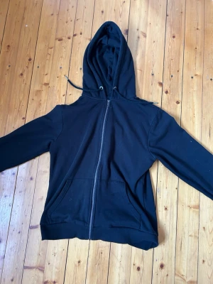 Svart hoodie med dragkedja - En klassisk svart hoodie med dragkedja framtill och huva med dragsko. Från lager 157. Hoodien har två fickor på magen och ribbade muddar vid ärmslut och nederkant. Perfekt för en avslappnad och stilren look. Storlek S.