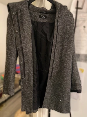 Lång kappa från ONLY XS Xsmall - Stilren grå kappa från ONLY med huva och dragkedja framtill. Kappan har ett mjukt, stickat yttertyg och svart foder på insidan. Långa ärmar och knappdetaljer längs dragkedjan ger en modern look. Defekter förekommer, se bild.