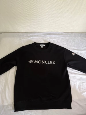 Svart sweatshirt från Moncler - Snygg svart sweatshirt från Moncler med vit broderad logga på bröstet och klassisk Moncler-patch på ärmen. Tröjan har rund halsringning och ribbade muddar vid ärmslut och nederkant. Perfekt för en clean och stilren look.