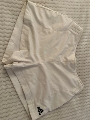 Vita sportiga shorts från Adidas - Säljer ett par vita sportiga shorts från Adidas med elastisk midja och diskret logga på ena benet. Shortsen har en enkel och clean design, perfekta för träning eller sommarens varma dagar. Använd ett par gånger i storlek M men passar som S