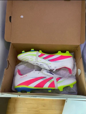Adidas Predator vita fotbollsskor - Säljer ett par Adidas Predator fotbollsskor i vitt med neonrosa och neongula detaljer samt blåa inslag. Skorna har klassisk snörning, låg profil och dobbar för spel på gräs. Snygg och modern design som sticker ut på planen.