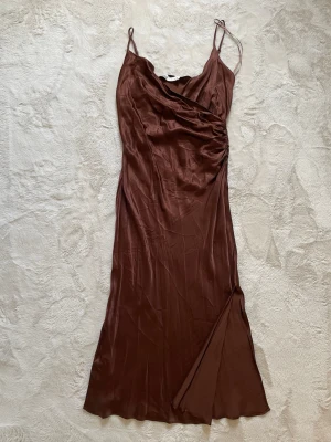 Brun långklänning med slits L ZARA - Snygg brun långklänning i silkesliknande material med tunna axelband och draperad detalj vid bysten. Klänningen har en slits framtill som ger en extra touch. Perfekt för sommarkvällar eller speciella tillfällen.