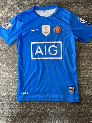 Blå Manchester United Nike tröja - Snygg blå Manchester United matchtröja från Dri-Fit med AIG som sponsortryck på bröstet. Tröjan har FIFA World Champions 2008-märke, klubbmärke och Nike-logga framtill. Korta ärmar och rund halsringning med vit kant. Perfekt för fotbollsfans! CR7. Bra skick.storlek 164