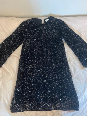 Svart glittrig paljettklänning H&M - Säljer en svart kort klänning från H&M täckt med glittriga paljetter. Klänningen har lång ärm och rund halsringning, perfekt för dig som vill sticka ut. Dragkedja i sidan för enkel påklädning.