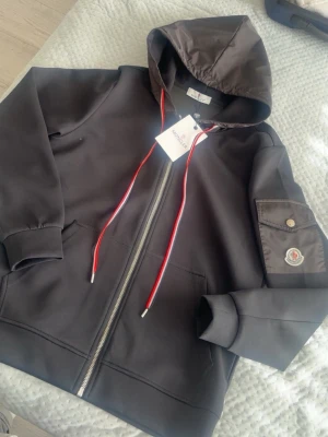 Svart hoodie med dragkedja från Moncler - Snygg svart hoodie från Moncler med dragkedja framtill och huva med röda och vita dragsnören. Ärmen har en ficka med lock och Moncler-logga. Tillverkad i mjukt material och har ribbade muddar vid ärmslut och nederkant.